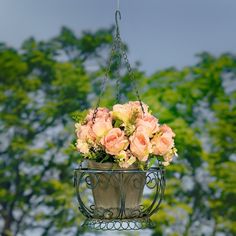 pink roses are in a hanging basket on a metal stand with trees in the background