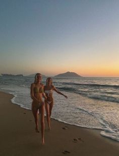 two women in bikinis running on the beach at sunset with footprints in the sand