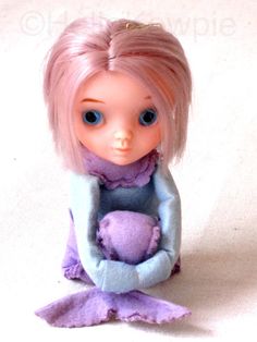 a doll with pink hair sitting on the floor holding a stuffed animal in her hands