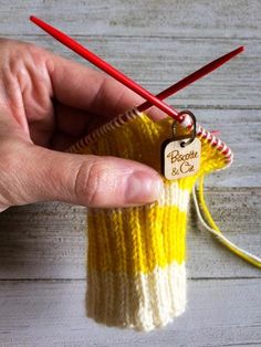 a hand holding two knitting needles next to a yellow and white knitted bag