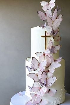 a three tiered cake with pink butterflies on it and a cross at the top