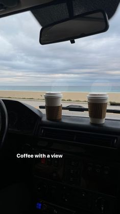 two cups of coffee sit on the dashboard of a car in front of an ocean