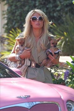 a woman holding two small dogs in her arms while standing next to a pink car