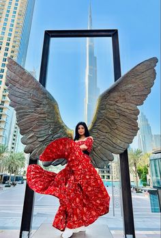 a woman in a long red dress with large wings standing next to a tall building