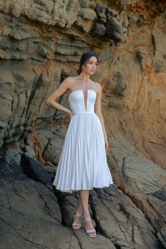 a woman in a white dress standing on rocks near the ocean with her hands on her hips