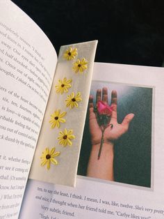 an open book with yellow flowers on it and a hand holding a flower in the middle