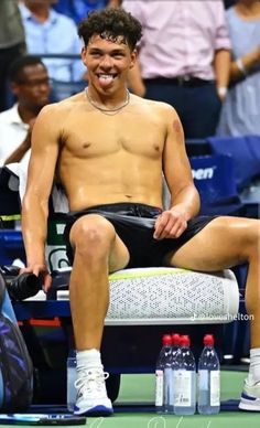 a shirtless tennis player sitting on a bench with water bottles in front of him