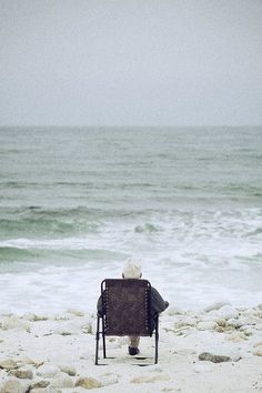 an old man sitting in a chair on the beach looking out at the ocean and waves