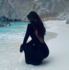 a woman sitting on top of a sandy beach next to the ocean in black dress