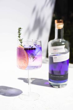 a purple drink sitting next to a bottle on top of a white tablecloth with a sprig of rosemary in it