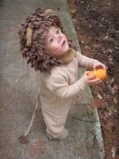 a toddler dressed up as a lion eating an orange on a sidewalk in the woods