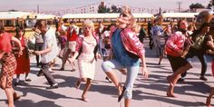 a group of people are dancing on the sidewalk in front of school buses and buildings