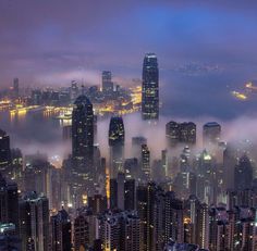 an aerial view of the city lights and skyscrapers at night with fog in the air