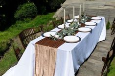 a long table with white plates and candles on it is set for an outdoor dinner