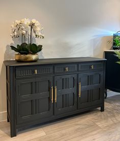 a black buffet with gold handles and flowers on top in a white walled room next to a planter