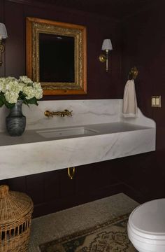 a white sink sitting under a mirror next to a toilet and a vase with flowers in it