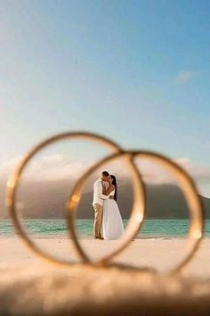 a bride and groom standing on the beach with their wedding rings in front of them