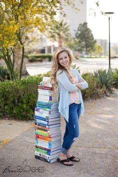 a woman standing next to a stack of books in front of some bushes and trees