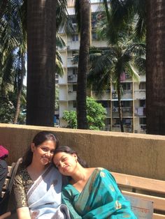 two women sitting on a bench next to each other in front of some palm trees