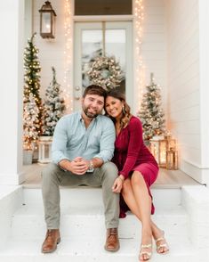 a man and woman sitting on the front steps of a house with christmas trees in the background