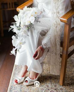 a bride sitting in a chair with her wedding bouquet on her hand and shoes off to the side