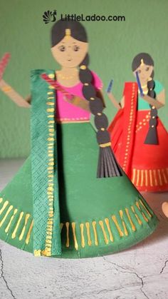 two paper dolls are standing next to each other on a table, one is wearing a green dress and the other is holding a fan