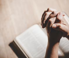 two hands clasped over an open book on a wooden table with a pen in it