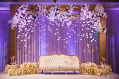 a couch sitting under a purple flower covered stage with flowers hanging from it's ceiling