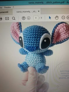 a hand holding a small blue and pink crocheted stitched stuffed animal in front of a computer screen