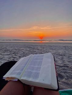 an open book sitting on top of a person's lap near the ocean at sunset