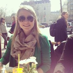 a woman sitting at a table with drinks in front of her and people walking by