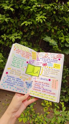 a hand holding an open notebook with writing and stickers on the pages in front of some bushes