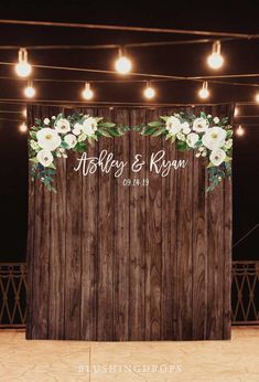 a wooden backdrop with white flowers and greenery on the top, along with string lights