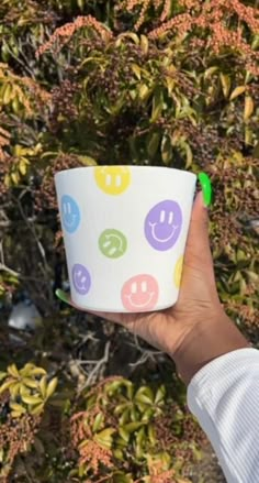 a person holding up a cup with smiley faces on it in front of some bushes