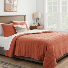 a bed with an orange comforter and pillows in a white room next to a window