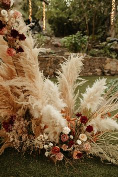 an arrangement of dried flowers and grasses on the grass in front of a stone wall