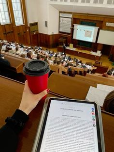 a person holding a coffee cup and tablet in front of an auditorium full of people