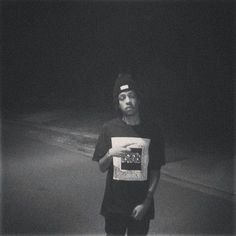 a man standing in the dark with his skateboard under his arm and wearing a t - shirt