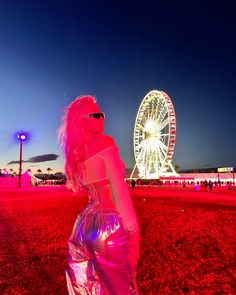 a woman standing in front of a ferris wheel at night with pink hair and sunglasses