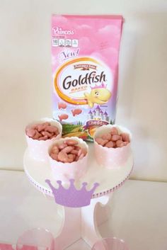 three pink cups filled with food next to a bag of goldfish crackers on a cake stand