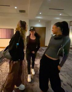 three young women are talking in an empty room with carpeted floors and large windows