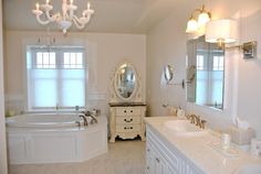 a white bathroom with two sinks and a bathtub in the corner, along with a chandelier