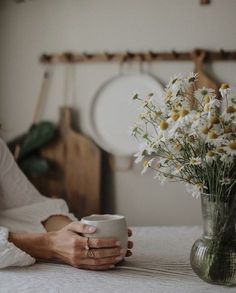 a woman sitting at a table holding a cup with flowers in the vase next to her