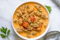 a white bowl filled with meat and tomato soup next to two spoons on a marble surface