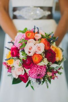 a woman holding a bouquet of flowers in her hands and wearing a white dress with red, pink, yellow and orange flowers on it