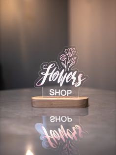 a glass sign that says flowers shop on top of a table with a candle in the background