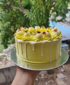 a hand holding a yellow cake with white icing and flowers on the top, in front of some trees