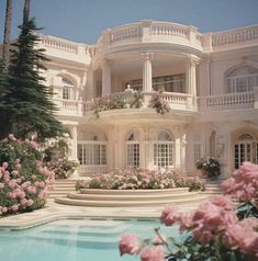 a large white house with pink flowers in the foreground and a pool surrounded by trees