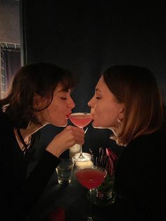 two women sitting at a table with drinks in front of their faces, one drinking from a wine glass