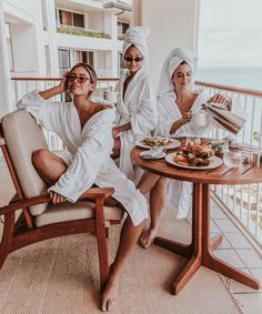 three women in white robes sitting at a table with food and drinks on the balcony